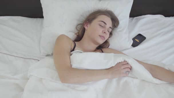 Ung vacker kvinna som ligger i sängen på morgonen. Flickan att stänga av väckarklockan på sin mobiltelefon och skarpt öppna ögonen. Flickan är sen till jobbet eller studien — Stockvideo