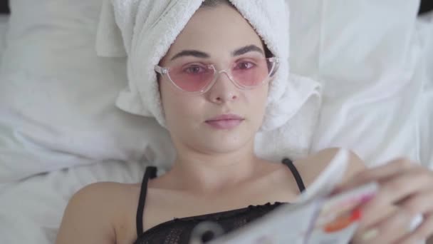 Linda mujer joven en gafas de color rosa y toalla en la cabeza acostada en la cama leyendo la revista, diario después de una ducha. Ocio en casa, día libre — Vídeos de Stock