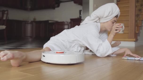 Bastante joven chica caucásica flexible después de una ducha en un albornoz sentado en cordel leyendo una revista bebe café o té en casa. Spinning robot aspiradora sala de limpieza . — Vídeos de Stock