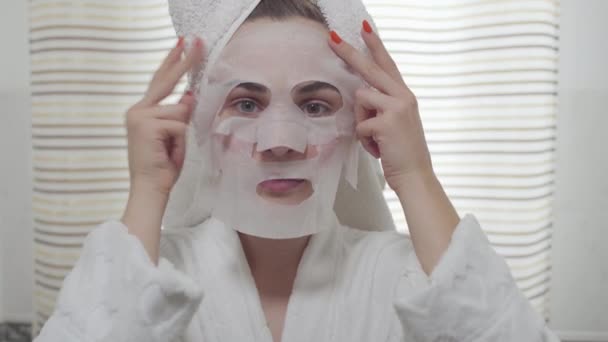 Kameraya bakan neşeli genç kadın banyoda yüzüne çarşaf maskesi uyguluyor. Farklı renkli gözleri ile sevimli kız. Cilt bakımı, güzellik konsepti. Gerçek insanlar serisi. — Stok video