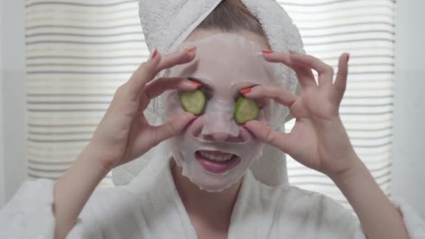 Attraktiv ung kvinna äter gurka med arket masken på hennes ansikte i Bad rums. Söt tjej med olika färgade ögon. Hudvård, skönhetskoncept. Real People-serien. — Stockvideo