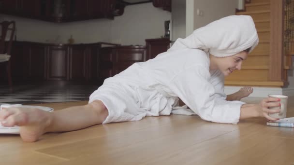 Cheerful young flexible caucasian girl after a shower in a bathrobe sitting on twine reading a magazine drinks coffee or tea at home. Spinning robot vacuum cleaner cleaning room. — Stock Video
