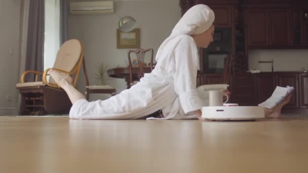 Cute young flexible caucasian girl after a shower in a bathrobe sitting on twine reading a magazine drinks coffee or tea at home. Spinning robot vacuum cleaner cleaning room. — Stock Footage Cute young flexible caucasian girl after a shower in a bathrobe sitting on twine reading a magazine drinks coffee or tea at home. Spinning robot vacuum cleaner cleaning room. Video Clip