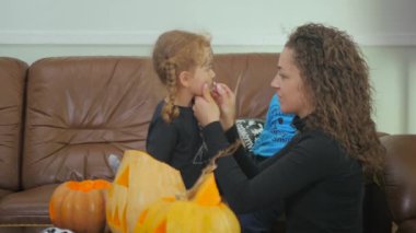 Kıvırcık kızı Cadılar Bayramı partisine hazırlayan kadının videosu. Kadın yüzüne makyaj yapar, çocuk balonlu.