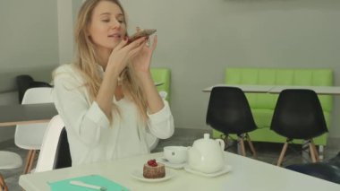 Kafedeki masada oturan genç ve tatlı bir kadın. Masada beyaz çaydanlık, fincan ve meyveli tatlı kek var. Genç ve güzel bir kadın akıllı telefonuyla fotoğraf çekiyor. Kadın elindeki aleti çevirir ve diğer taraftan bir fotoğraf çeker..