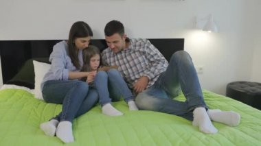 Mutlu bir aile yatak odasında büyük yeşil bir yatakta yatıyor. Küçük tatlı kız elinde bir tablet tutuyor. Gençler tablete bakıyor, ekrana dokunuyor ve gülümsüyor. Genç aile birlikte rahatlıyor ve mutlular..