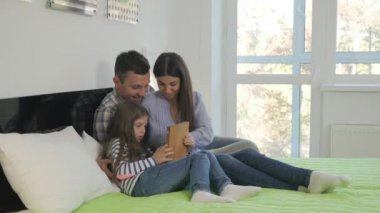 Mutlu bir aile yatak odasında büyük bir yatakta yatıyor. Küçük tatlı kız elinde bir tablet tutuyor. Gençler tablete bakıyor, ekrana dokunuyor ve gülümsüyor. Yakışıklı genç bayan saçını düzeltiyor..