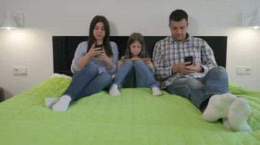 Genç bir aile, yatak odasındaki yeşil bir yatağa uzanıp aletlerini kullanıyor. Gülümseyen kadın akıllı telefonunu adama gösteriyor. Küçük kız ve adam ekrana bakıyor. Kadın kocasına bir şeyler söylüyor..
