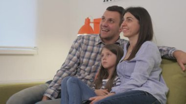 Genç mutlu bir aile yeşil bir kanepede oturuyor ve televizyona bakıyor. Küçük kız ebeveynlerinin ortasında oturuyor. Genç bir kadın uzaktan kumandayla televizyondaki kanalları değiştiriyor. Ebeveynler kızlarına bakıp gülümserken kadın televizyonu değiştiriyor..