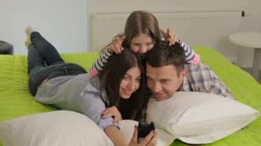 Genç bir kadın ve çekici bir adam yeşil yatakta beyaz bir piilovda yatıyor. Sırtlarında tatlı bir kız yatıyor. Mutlu aile akıllı telefondan selfie çeker. Küçük kız ailesinin kafasında iki parmağını gösteriyor..
