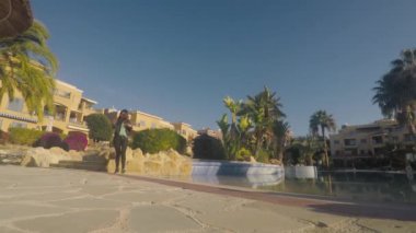 Siyah ceketli, akıllı telefonu elinde tutan ve yüzme havuzundan kameraya doğru yürüyen genç bir esmer. Güzel bir kız akıllı telefondan video çeker ve tatil köyünü çekmek için geri döner..