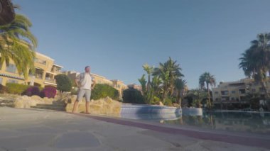 Genç sporcu ellerini ortaya koyar, bacaklarını omuz genişliğine koyar ve giyinik gözlerle güneşe bakar. Adam bacaklarını birleştiriyor ve havuzun yanında çömelmeye başlıyor..