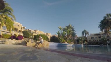 Komik adam yüzme havuzunun yanında görünmez rakibinle dövüşecek pozisyonda duruyor. Adam pozisyonunu değiştiriyor, arkasını dönmeye çalışıyor ve su sıçratmak için bacağını kaldırıyor. Adam ellerini farklı taraflara koyuyor..