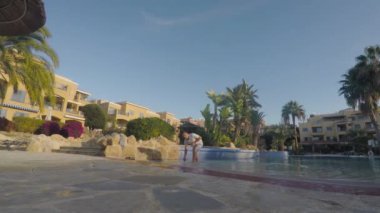 Tişörtlü genç adam tatil köyü bölgesinde yüzme havuzunda suyun içinde duruyor. Çekici adam kameraya su sıçratmaya çalışıyor. Adam suya su sıçrattı, kameraya baktı ve başparmağını gösterdi..