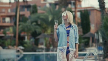 Gözlüklü ve bikinili tatlı sarışın yüzme havuzunun yanında duruyor ve elinde şampanya bardağıyla duruyor. Güzel kız gülümsüyor, elini ortaya koyuyor ve mavi bir yüzme havuzuna bakıyor. Kız kameraya bakıyor, gülümsüyor ve poz veriyor..