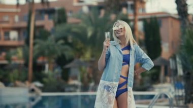 Gözlüklü ve bikinili güzel sarışın yüzme havuzunun yanında duruyor ve elinde şampanya bardağıyla duruyor. Güzel kız gülümsüyor, elini ortaya koyuyor ve kameraya bakıyor. Gülümseyen ve poz veren tatlı kız..