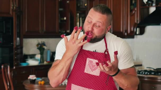 有纹身的肌肉发达的帅哥正在吃着手指上的覆盆子 快乐的人一个接一个地拿走手中的浆果 年轻有趣的人在厨房吃覆盆子 — 图库视频影像