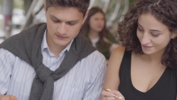 Portrait d'un couple heureux mangeant du désert assis dans un café à l'extérieur. Un jeune homme et une jeune femme qui s'amusent se retrouvent assis ensemble à savourer leur repas. Concept de date au restaurant . — Video