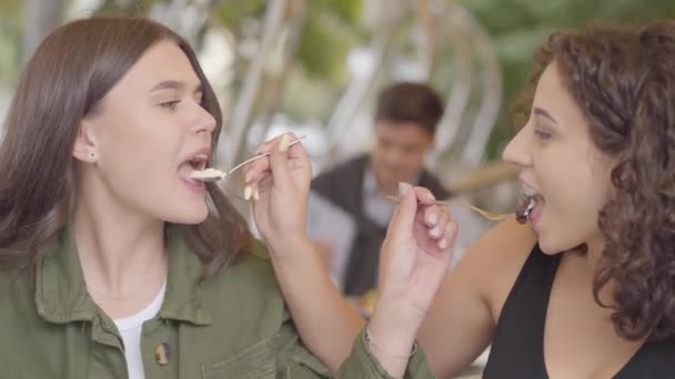 Retrato de dos novias alimentándose mutuamente con un pastel sentado en la cafetería al aire libre. Chicas jóvenes juntas disfrutando de su comida. Ocio exterior — Vídeo de stock