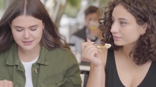 Retrato de dos novias comiendo un pastel sentado en la cafetería al aire libre. Chicas jóvenes juntas disfrutando de su comida hablando y riendo. Ocio al aire libre, relación amistosa — Vídeo de stock