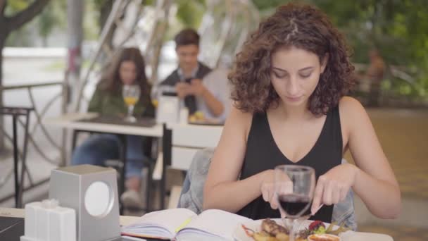 Jonge mooie krullend vrouw eten lekker vlees eten in straat Cafe. Het meisje geniet van haar maaltijd ontspannen buiten. De dame snijden kotelet met vork en mes. Vrije tijd buitenshuis — Stockvideo