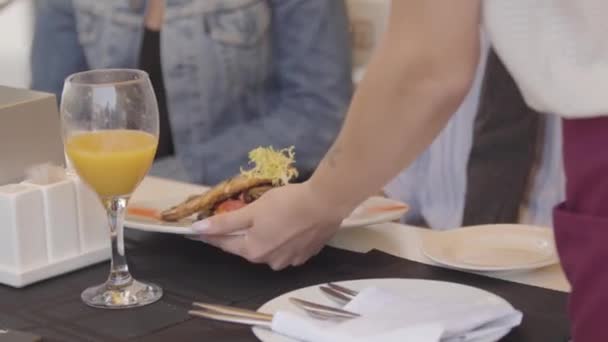 Mains de jeune serveuse avec tatouage mettant du poisson et de la viande sur la table devant les visiteurs masculins et féminins. Le couple heureux profitant de leur dîner relaxant dans un café. Loisirs ensemble, concept de date — Video