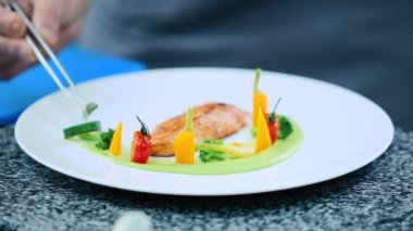 Soslu tabak, balık filetosu ve brokoli masada duruyor. Aşçı balık filetosunu kuşkonmazla süslüyor.