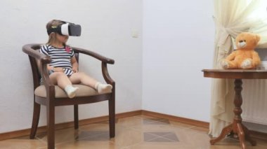 Sanal gözlüklü şirin küçük kız sandalyede oturuyor ve etrafa bakıyor. Küçük çocuk evde sanal gerçeklik gözlüğü kullanıyor. Kız gözlüğünü kafasından çıkarır ve gülümser. Gözlüklü çocuk şaşırdı..