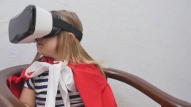 Cute little girl in red cloak and in virtual glasses is sitting at the chair and looks around. Little kid uses a virtual reality glasses at home. Girl takes off the glasses from head and smile. The kid in surprise from image in glasses.
