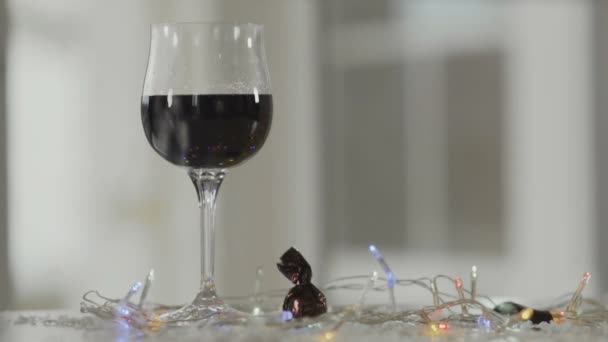 装飾的な雪は赤ワインとカラフルなクリスマスライトでワイングラスの近くに落ちます — ストック動画