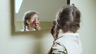 Küçük kız banyoda aynanın önünde duruyor ve kendisi için makyaj yapıyor. Sevimli çocuk, yetişkin annesi gibi olur. Kız pudra fırçasıyla yanaklarını boyuyor..