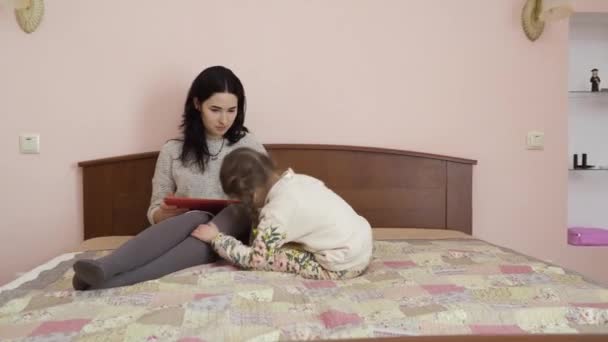 妈妈和小女孩坐在家里的床上拥抱着 漂亮的女人和可爱的女孩表现出彼此的感情 这个女人膝盖上抱着数码平板电脑 — 图库视频影像