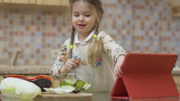 可爱的小女孩在厨房里学习烹调沙拉 漂亮的孩子把沙拉和勺子混合在一个碗里 然后在数码平板电脑上看电影 可爱的女孩尝她新鲜的沙拉 Prores Codec — 图库视频影像