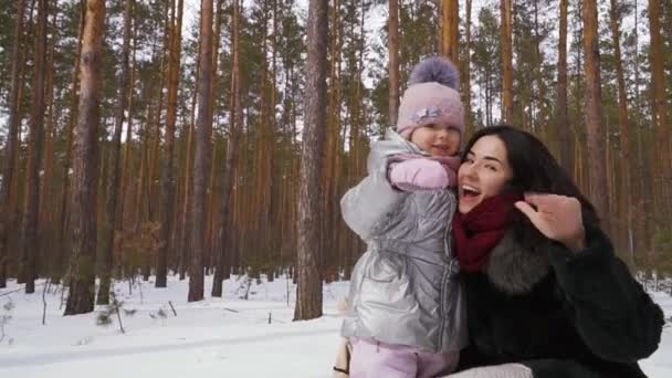 幼い娘を持つ若い女性が冬の森を歩く 家族は屋外で一緒に余暇を過ごします カメラを見て手を振ってるスローモーションだコーデックを証明する — ストック動画