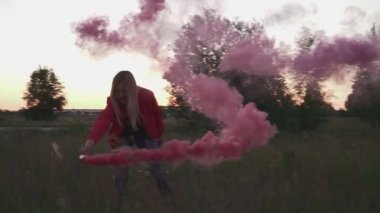 Pembe ceketli sarışın kız açık havada dairesel hareketlerle renkli sis bombası püskürtüyor. Video ve fotoğraf çekimleri için özel dekorasyon kullanılıyor. Dışarıda vakit geçirmek.