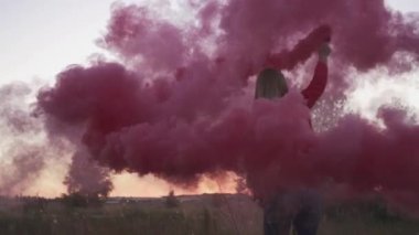 Sarışın genç bayan mor duman sütunu etrafında dönüyor. Kadın açık havada dairesel hareketlerle renkli sis bombası püskürtüyor. Video ve fotosezi için özel dekorasyon kullanılıyor. Dışarıda vakit geçirmek.