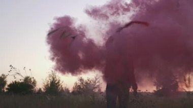 Sarışın genç bayan mor duman sütunu etrafında dönüyor. Kadın açık havada dairesel hareketlerle renkli sis bombası püskürtüyor. Video ve fotosezi için özel dekorasyon kullanılıyor. Dışarıda vakit geçirmek.