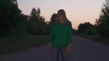 Yeşil kazaklı sarışın kız yolun kenarında duruyor. Bayan saçını düzeltir ve yavaşça dans etmeye başlar. Dışarıda vakit geçirmek.
