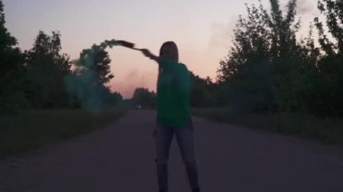 Yeşil ceketli sarışın kız açık havada dairesel hareketlerle renkli sis bombası püskürtüyor. Video ve fotoğraf çekimleri için özel dekorasyon kullanılıyor. Akşam vakti dışarıda vakit geçirmek.