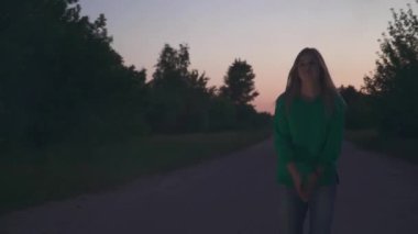 Yeşil kazaklı sarışın kız yol boyunca yürüyor ve saçını düzeltiyor. Bayan kameraya bakar ve geçer.