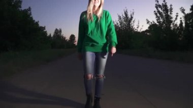 Yeşil kazaklı sarışın kız yol boyunca yürüyor ve saçını düzeltiyor. Bayan kameraya bakar ve geçer.