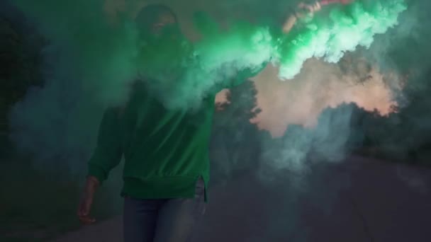 緑色のジャケットのブロンドの女の子は オープンエアの円形の動きで色の煙爆弾を噴霧しています ビデオや写真セッションのための特別な装飾を使用してください 夕方の時間帯に屋外で過ごす — ストック動画