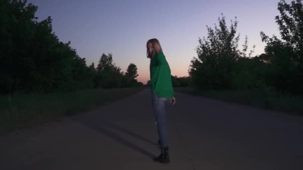 若い女性は手の波で彼女に従ってください 緑のプルオーバーのブロンドの女の子が道に沿って走っています 夕方の時間帯に屋外で過ごす — ストック動画