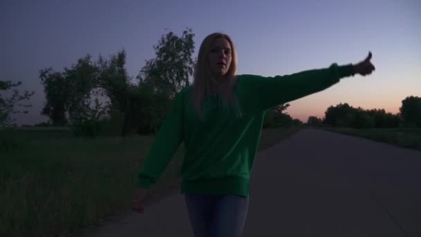 若い女性は手の波で彼女に従ってください 緑のプルオーバーのブロンドの女の子が道に沿って走っています 夕方の時間帯に屋外で過ごす — ストック動画