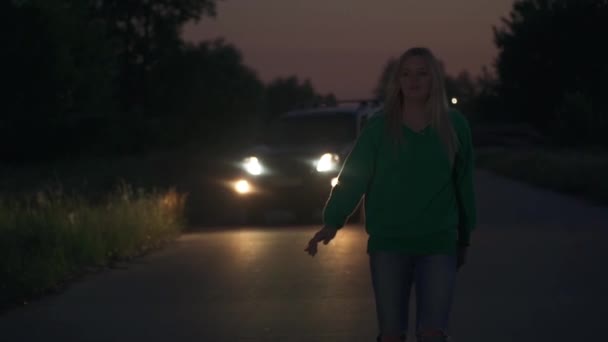 傍晚时分 开着前灯的黑色轿车尾随着穿着绿色夹克的金发姑娘 这个女孩在搭便车 — 图库视频影像