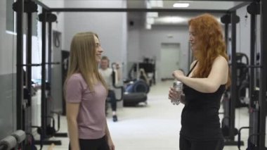 Spor salonunda kızıl saçlı kadına bir şişe su veren pozitif kadın eğitmen. Kişisel antrenörle çalışan kız. Spor kavramı, aktif yaşam tarzı, sağlık