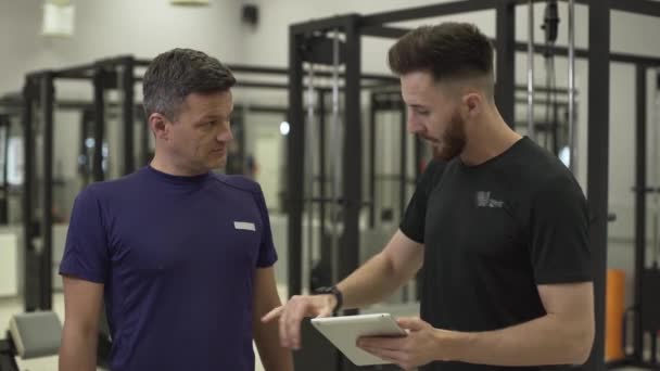 Elinde bir tablet ile profesyonel fitness eğitmeni bir sporcu için eğitim uçağı açıklayan. Adam spor salonunda vücudunu geliştirecek. Spor kavramı, aktif yaşam tarzı, sağlık — Stok video