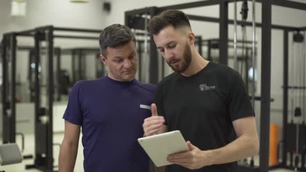 Guapo entrenador de fitness profesional con una tableta en las manos explicando el avión de entrenamiento para un deportista. El hombre va a mejorar su cuerpo en el gimnasio. Concepto de deporte, estilo de vida activo — Vídeos de Stock