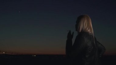 Deri ceketli sarışın kadın havaalanının arka planında sigara içiyor. Uçağın gökyüzü ışığı yanıp sönüyor. Akşamın geç saatlerinde vakit geçirmek