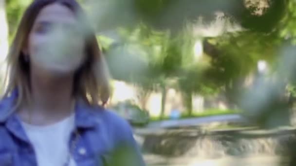 若い美しい女性が夏の公園を歩いている きれいなブロンドは木の花を楽しむ 魅力的な女の子でデニムジャケットリラックス屋外 — ストック動画